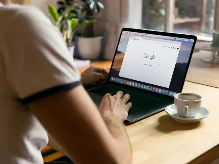 Person browsing Google on a laptop at a wooden table with a cup of coffee in a cozy workspace